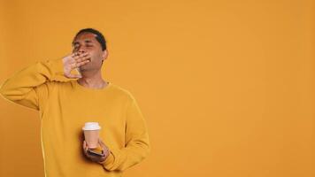 Indian man enjoying coffee from disposable paper cup and send text messages on smartphone. Person drinking hot beverage and chatting with friends on mobile phone, studio background, camera A video