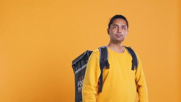 Portrait of confident indian man holding thermal backpack, crossing arms, studio background. Radiant nonchalant delivery person prepared to deliver takeaway food to customers, camera B video