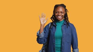 retrato do picador menina segurando mochila, acenando mão, gesticulando, isolado sobre amarelo estúdio fundo. alegre africano americano aluna com escola mochila fazendo cumprimento mão gesto, Câmera uma video