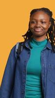 Vertical Portrait of happy woman waving hand, saluting, gesturing, isolated over yellow studio background. Cheerful BIPOC teenager with school rucksack doing greeting hand gesture, camera A video