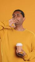 Indian man enjoying coffee from disposable paper cup and send text messages on smartphone. Person drinking hot beverage and chatting with friends on mobile phone, studio background, camera A video