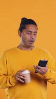 Indian man enjoying coffee from disposable paper cup and send text messages on smartphone. Person drinking hot beverage and chatting with friends on mobile phone, studio background, camera B video