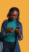 Vertical African american teenager adding payment method on website, isolated over studio background. Young girl wearing headphones, doing internet shopping, typing credit card information on phone, camera B video