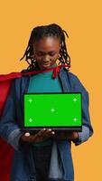 Vertical Portrait of jolly teenager acting as superhero presenting green screen laptop, isolated over studio background. Cheerful BIPOC person posing as hero showing mockup notebook features, camera B video