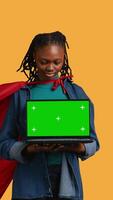 Vertical Portrait of BIPOC woman portraying superhero with cape presenting isolated screen laptop, studio background. African american young girl posing as hero talking, showing chroma key notebook, camera B video