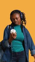 Vertical Young girl with backpack and headphones yawning, feeling tired, drinking coffee to wake up. Sleepy woman using caffeinated beverage to gain energy, lacking sleep, studio background, camera B video