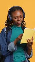 vertical joven niña vistiendo auriculares leyendo interesante libro, disfrutando pasatiempo, siendo entretenido, aislado terminado estudio antecedentes. optimista africano americano mujer con novela en brazos relajante, cámara un video