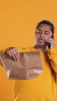 Man packing food in thermal backpack, fulfilling clients orders and answering phone ringing, studio backdrop. Indian person doing delivery to customers, discussing with them on cellphone, camera B video