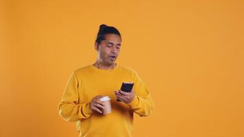 Indian man enjoying coffee from disposable paper cup and send text messages on smartphone. Person drinking hot beverage and chatting with friends on mobile phone, studio background, camera B video