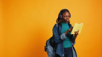 Young girl wearing headphones reading interesting book, enjoying hobby, being entertained, isolated over studio background. Upbeat african american woman with novel in arms relaxing, camera B video