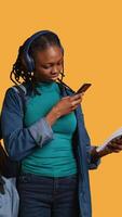 Vertical African american woman taking picture of school homework to send to colleague, listening music, studio background. Student photographing papers with information needed for exam, camera B video