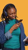 Vertical African american woman taking picture of school homework to send to colleague, listening music, studio background. Student photographing papers with information needed for exam, camera A video