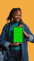 Vertical Smiling african american woman presenting tablet with green screen display, isolated over studio background. Cheerful BIPOC person creating promotion with chroma key device, camera B video