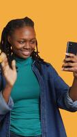 Vertical Cheerful woman saluting friends during teleconference meeting using smartphone, studio background. Teenager having fun catching up with school colleagues during videocall, camera A video