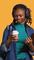 Vertical African american teenager enjoying coffee while holding clipboard. Young person doing productivity tasks, drinking caffeinated beverage, isolated over studio background, camera A video