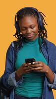 Vertical Happy person having fun, dancing on rhythm, enjoying songs during leisure time. Cheerful BIPOC woman doing funky movements on music playing on headphones, studio background, camera A video