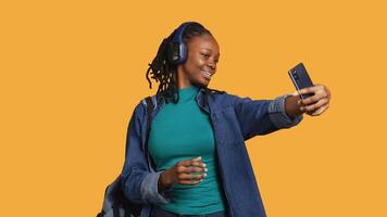 vrolijk tiener gebruik makend van mobiele telefoon naar nemen selfies aan het doen zege hand- teken. glimlachen bipoc meisje nemen afbeeldingen gebruik makend van mobiel telefoon, aan het doen zege gebaren, luisteren muziek, studio achtergrond, camera een video