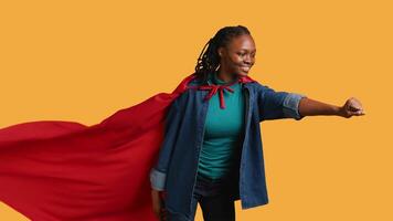 Happy girl portraying superhero flying with cape fluttering in wind, isolated over studio background. Upbeat heroic african american woman posing as hero in costume, camera B video