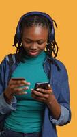 vertical africano americano adolescente adicionando Forma de pagamento método em local na rede Internet, isolado sobre estúdio fundo. jovem menina vestindo fones de ouvido, fazendo Internet compras, digitando crédito cartão em formação em telefone, Câmera uma video