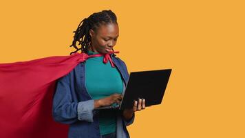 Happy african american young girl wearing superhero costume for Halloween working on laptop, studio background. Jolly teenager dressed as comic book hero typing on notebook keyboard, camera A video