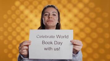 Portrait of happy woman holding placard with world book day message written on it, isolated over studio background. Geek promoting importance of reading during 23th April global event, camera B video