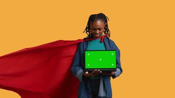 Portrait of jolly teenager acting as superhero presenting green screen laptop, isolated over studio background. Cheerful BIPOC person posing as hero showing mockup notebook features, camera B video