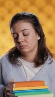 Vertical Exhausted woman yawning, holding heavy stack of books needed for school exam, studio background. Student yearning for sleep, tired of learning from academic textbooks, camera B video