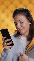 Vertical Smiling woman reading messages on smartphone while holding stack of books, isolated over studio background. Cheerful person with pile of academic textbooks in arms texting friends on phone, camera B video