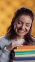 verticaal portret van gelukkig vrouw met stack van boeken in handen tonen duimen omhoog, studio achtergrond. vreugdevol boekenwurm Holding stapel van romans, gevoel vrolijk, aan het doen positief hand- gebaren, camera een video