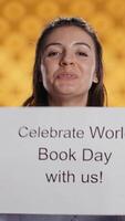 Vertical Portrait of happy woman holding placard with world book day message written on it, isolated over studio background. Geek promoting importance of reading during 23th April global event, camera B video