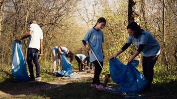 Mom and child collecting rubbish from the forest area using a long claw tool and garbage disposal bags. Little girl picking up trash and plastic waste with her mother, ecological justice. Camera B. video