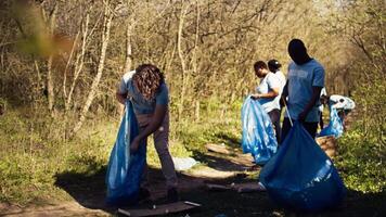 Diverse men volunteers pick up rubbish and plastic trash with tongs, working to combat illegal dumping and preserve natural forest environment. Activists volunteering for litter cleanup. Camera B. video