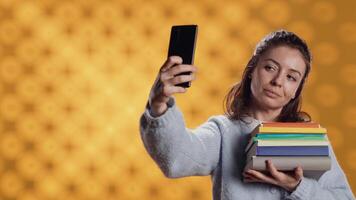 sonriente mujer participación pila de libros, disfrutando leyendo pasatiempo, tomando selfies con Teléfono móvil. alegre dama con apilar de novelas en brazos haciendo imágenes con móvil teléfono, estudio fondo, cámara un video