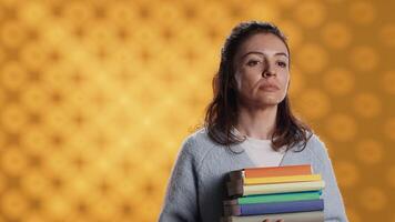 retrato de optimista mujer participación pila de libros, disfrutando leyendo pasatiempo para entretenimiento propósitos. radiante ratón de biblioteca con apilar de novelas en brazos disfrutando ocio tiempo, estudio fondo, cámara un video