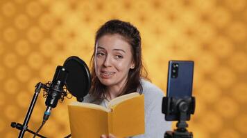 Media star filming vlog at work, reading book to record audiobook, studio background. Voice actor doing content creation while producing digital recording on novel using microphone, camera A video