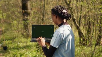 African american girl cleaning up forest and using isolated laptop display, taking part in a litter cleanup activity to protect the ecosystem and wildlife. Nature preservation concept. Camera A. video