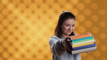 Portrait of friendly woman offering stack of textbooks useful for school exam, isolated over studio background. Merry person giving pile of books, recommending them for university assessment, camera A video