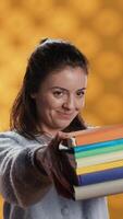 Vertical Portrait of friendly woman offering stack of textbooks useful for school exam, isolated over studio background. Merry person giving pile of books, recommending them for university assessment, camera A video