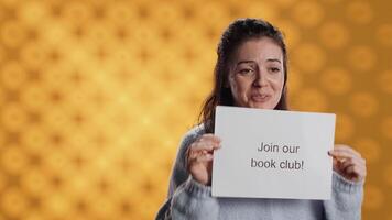 Cheerful book club president holds message urging people to join them, talking about importance of lecture, studio background. Smiling woman inviting bookworms to enlist in her association, camera A video