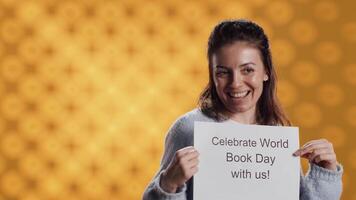 Portrait of happy woman holding placard with world book day message written on it, isolated over studio background. Geek promoting importance of reading during 23th April global event, camera A video