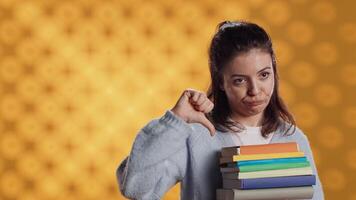 portret van vrouw met pruilen uitdrukking Holding stapel van boeken, tonen afkeuring van lezing hobby. sulky dame met stack van romans aan het doen duimen naar beneden hand- gebaren, studio achtergrond, camera een video