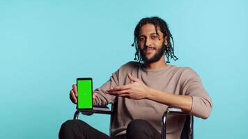 middle eastern man in wheelchair pointing towards green screen mobile phone. Person with paraplegia presenting chroma key cellphone, isolated over blue studio background video