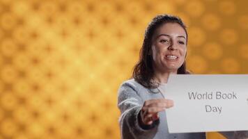 Smiling book club member holding placard with world book day message, isolated over studio background. Radiant bookworm recording promoting literacy importance, camera A video