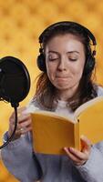 Vertical Voice actor reads book while enjoying coffee, recording audiobook using microphone, studio background. Woman using mic and headphones to produce digital recording of novel, drinking beverage, camera B video