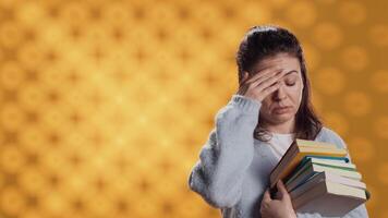 Woman with worried facial expression feeling migraine pain, carrying books for school exam, studio background. Stressed student with textbooks suffering headache from too much homework, camera A video