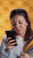 Vertical Smiling woman reading messages on smartphone while holding stack of books, isolated over studio background. Cheerful person with pile of academic textbooks in arms texting friends on phone, camera A video