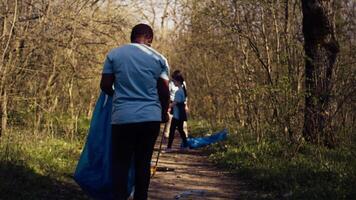 Diverse activists group using claw tools to grab trash and plastic, storing it in garbage disposal bags. People gathering to collect rubbish and recycling for sustainable lifestyle. Camera B. video