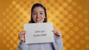 Cheerful book club president holds message urging people to join them, talking about importance of lecture, studio background. Smiling woman inviting bookworms to enlist in her association, camera B video