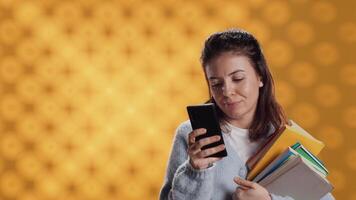 sonriente mujer leyendo mensajes en teléfono inteligente mientras participación apilar de libros, aislado terminado estudio antecedentes. alegre persona con pila de académico libros de texto en brazos mensajes de texto amigos en teléfono, cámara un video