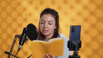 influenciador filmando lendo livro com Smartphone em tripé, criando áudio-livro, estúdio fundo. mulher vlogging com Móvel telefone enquanto produzindo digital gravação em romance usando microfone, Câmera uma video